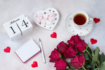 a cup of tea, a bouquet of red roses, heart-shaped marshmallows, satin hearts, a notebook, a pen and a calendar date February 14