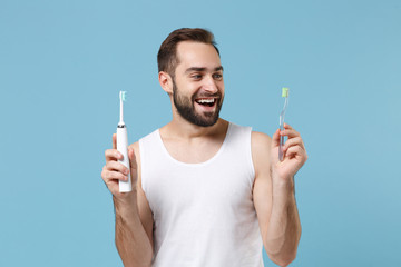 Bearded young man 20s years old in white shirt hold choice electric brushes isolated on blue pastel...