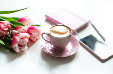 Pink notebook and cup of cappuccino coffee with pink tulips on white background. Pink writing set with cappuccino. Woman's workplace in pink. Flat lay. Top view. Copy space