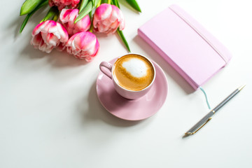 Pink notebook and cup of cappuccino coffee with pink tulips on white background. Pink writing set with cappuccino. Woman's workplace in pink. Flat lay. Top view. Copy space