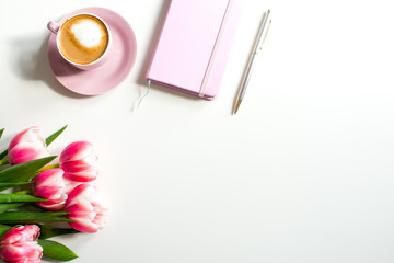 Pink notebook and cup of cappuccino coffee with pink tulips on white background. Pink writing set with cappuccino. Woman's workplace in pink. Flat lay. Top view. Copy space