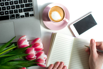 Young woman hands hold opened notebook pages. Morning coffee mug for breakfast, empty notebook, pencil and pink tulips flowers on white table top view in flat lay style. Woman working desk.