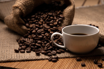 Coffee beans spill out of the bag. Nearby is a cup of coffee. Side view. Great postcard.
