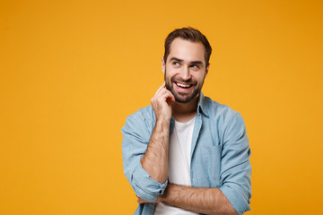 Smiling pensive young bearded man in casual blue shirt posing isolated on yellow orange background...