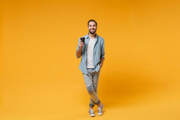 Fototapeta premium Smiling young man in casual blue shirt posing isolated on yellow orange wall background studio portrait. People sincere emotions lifestyle concept. Mock up copy space. Hold paper cup of coffee or tea.