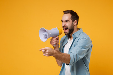 Crazy young man in casual blue shirt posing isolated on yellow orange wall background studio portrait. People lifestyle concept. Mock up copy space. Screaming in megaphone pointing index finger aside.