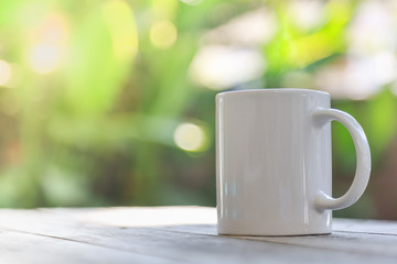 Close up of white mug cup of hot coffee on wooden table and bokeh green leaf nature under sunlight as background.