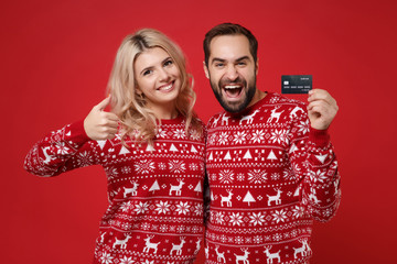 Funny young couple guy girl in Christmas sweaters posing isolated on red background. Happy New Year 2020 celebration holiday party concept. Mock up copy space. Hold credit bank card, showing thumb up.