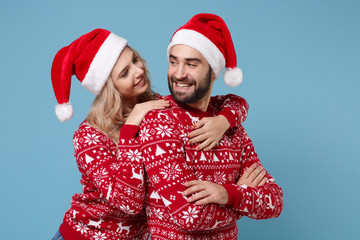 Pretty young couple guy girl in Christmas sweaters, Santa hat posing isolated on blue background. New Year 2020 celebration holiday party concept. Mock up copy space. Holding hands crossed, hugging.