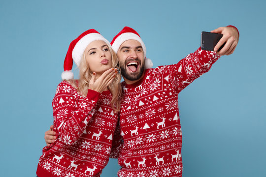 Young couple guy girl in Christmas sweater Santa hat posing isolated on blue background. New Year 2020 celebration concept. Mock up copy space. Doing selfie shot on mobile phone blowing send air kiss.