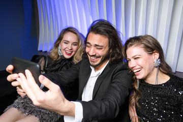 Laughing glamorous girls and elegant man with smartphone making selfie at party