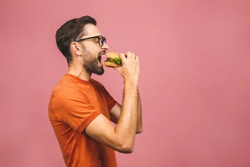 Gordijnen Young man holding a piece of hamburger. Student eats fast food. Burger is not helpful food. Very hungry guy. Diet concept. Isolated over pink background. © denis_vermenko