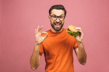Young man holding a piece of hamburger. Student eats fast food. Burger is not helpful food. Very hungry guy. Diet concept. Isolated over pink background. Ok sign.