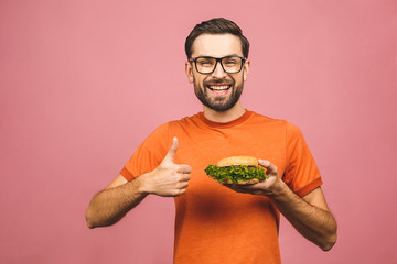 Young man holding a piece of hamburger. Student eats fast food. Burger is not helpful food. Very hungry guy. Diet concept. Isolated over pink background. Thumbs up.