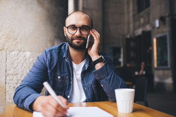 Smart man in glasses writing at table and speaking on smartphone