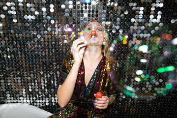 Young glamorous woman in glittering dress blowing soap bubbles at party
