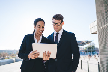 Collaborating diverse business people with tablet
Smiling black woman in suit showing tablet to male coworker in glasses while working in team and standing on background of cityscape