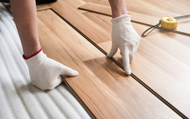 Installing laminated floor, detail on man hands in gloves fitting wooden tile, over white foam base...