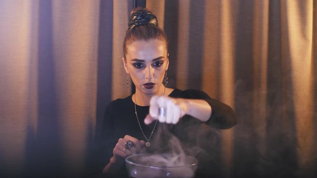 a witch over a bowl with a potion from which smoke comes out makes magical movements with her hands and looks into the camera
