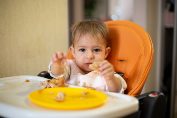 A cute baby in an orange child seat with a table, looks at camera and holds out a piece of cake. there are crumbs and an orange plate on the table. close-up, front view, soft focus, blur background