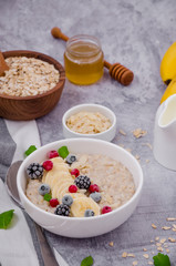 Oatmeal in a bowl with banana, frozen berries, almond petals, mint leaves and honey on a gray stone background. Tasty and healthy breakfast.