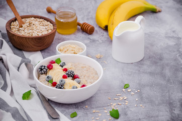 Oatmeal in a bowl with banana, frozen berries, almond petals, mint leaves and honey on a gray stone background. Tasty and healthy breakfast.