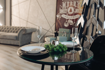 View over beautifully decorated round table with natural fir branch, candle, two flutes, plates against classic sofa in modern apartment.
