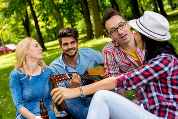 Group of young people in park enjoying time together, playing guitar and drinking beer