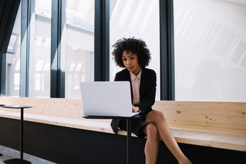 Afro american confident businesswoman dressed in formal wear concentrated on financial report typing on netbook and checking database sitting in office interior and using 4g wireless internet