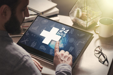 Man with laptop on his desk. He presses on the icon of medical care. Concept of the round-the-clock medical care.