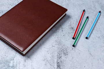 Red leather notepad with coloured pens on gray concrete background. Copy space for text. Minimalism blogging, freelancing concept.