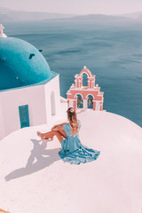 Young woman with blonde hair and blue dress in oia, santorini, greece with ocean view and churches - obrazy, fototapety, plakaty