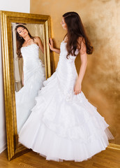 Beautiful dark haired woman in white bridal dress standing nearby mirrow and looking at it