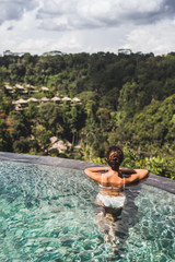 Woman in white swimsuit and sunglasses relaxing in luxury infinity pool with jungle view in Ubud, Bali. View from behind. Amazing Indonesian nature.