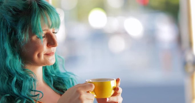 Attractive millennial woman with blue hair drinking from yellow cup, copy space bokeh