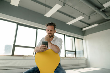 Mature businessman sitting on yellow chair in empty office using smartphone