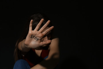 Abused young woman showing palm with word STOP against black background, focus on hand and space for text. Domestic violence concept