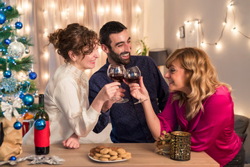 Friends having a toast with wine by Christmas tree in living room. Holidays preparation.