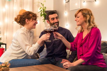 Friends having a toast with wine by Christmas tree in living room. Holidays preparation.