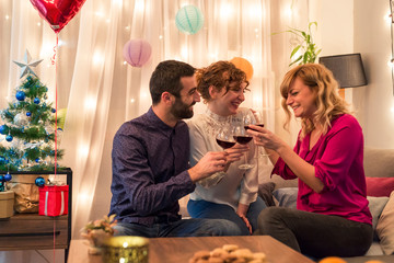 Friends having a toast with wine by Christmas tree in living room. Holidays preparation.