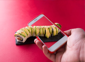 A hand holds a phone and photographs green sushi with avocados and this roll goes out of the scope of the phone. Close-up photo.