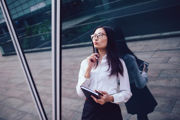 Thoughtful attractive female worker with notepad and mobile on street in front of business center