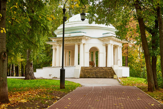 Stone white gazebo in the Park with an alley on an autumn day