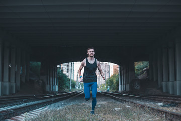 Healthy young man jogging in the city at night. Full length shot of male athlete running under bridge along the railroad trucks.