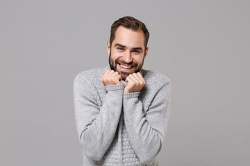 Smiling frozen young bearded man in gray sweater posing isolated on grey background studio portrait. Healthy fashion lifestyle people emotions cold season concept. Mock up copy space. Clenching fists.