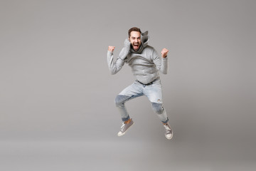 Fototapeta na wymiar Happy young bearded man in gray sweater, scarf posing isolated on grey background, studio portrait. Healthy fashion lifestyle, cold season concept. Mock up copy space. Jumping, doing winner gesture.