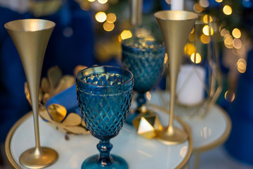 Blue glass and gold metallic holiday glasses, New Year's dinner, decor in blue and gold colors against the backdrop of a Christmas tree and lights