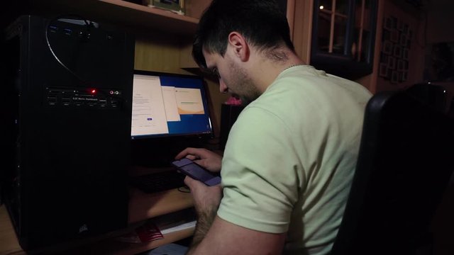 Brunette guy in green T-shirt sitting at table, near black system unit, boxes, typing on the keyboard, with smartphone code, password. In evening, in apartment, at home, in room.