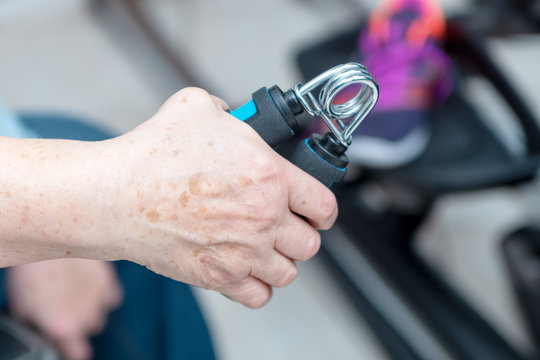 Handmuskeltrainer in der Hand einer Seniorin vor einem unscharf dargestellten Crosstrainer mit Sportschuh