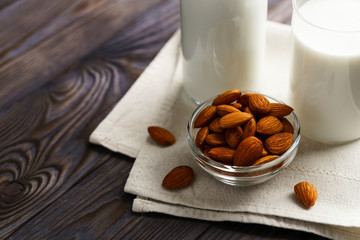 Close-up of almond milk in a glass mug and a refillable glass bottle, next to it lie almonds and a textile napkin on a wooden table.
